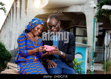 two happy adult businesspeople sitting on the street using a mobile phone. Stock Photo