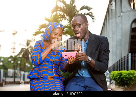 beautiful african couple sitting outdoors looking at cellphone smiling. Stock Photo