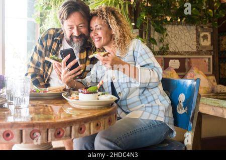 Adult caucasian couple man and woman in video conference call on mobile phone during lunch at cafe restaurant together. Cheerful happy hipster people Stock Photo