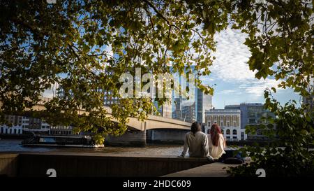 A young couple sits on the River Thames embankment next to London Bridge in the city centre as a tourist boat passes. Stock Photo