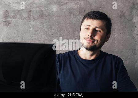 Handsome smiling bearded man looking at laptop screen against concrete wall in modern office Stock Photo
