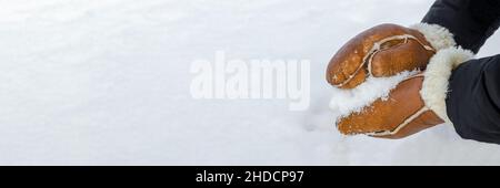 First snow, making snowballs in the park close-up. Hands in warm mittens make snow in winter. Brown leather mittens in the snow in winter. Stock Photo