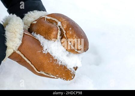 Woman in leather mittens rolls a snowball outdoors, close-up. Hands in warm mittens make snow in winter. Brown leather mittens in the snow in winter. Stock Photo