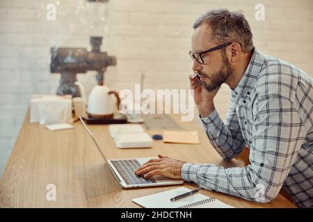 Bearded man talking on cellphone and using laptop in cafe Stock Photo