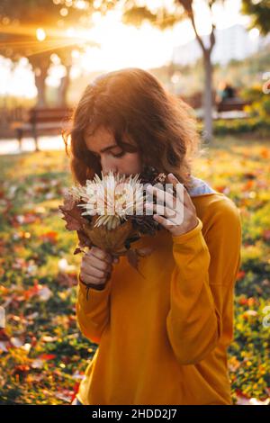 Curly young girl in yellow sweater on the grass with autumn bouquet of dry leaves and flowers Stock Photo