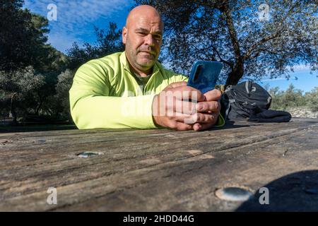 Low angle shot of a shaved-head Caucasian male sitting by a wooden table holding his smartphone Stock Photo