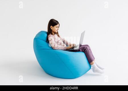 Child using laptop while resting on bean bag chair on white background Stock Photo