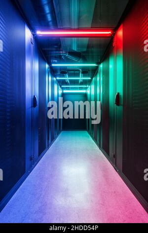 Cold aisle of an IT server room with perforated grille doors illuminated in color and large cooling pipes on the ceiling Stock Photo