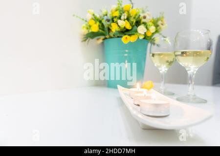 Romantic set up of burning candles, two  white wine glasses and blooming flowers in bucket on white table and wall in background Stock Photo