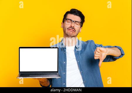 Upset crying caucasian modern guy with glasses, holding open laptop with blank white screen in hand, showing thumbs down, while standing against isolated orange background. Negative emotion Stock Photo