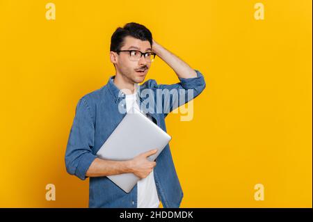 Puzzled discouraged guy in glasses and in a denim shirt, holding a laptop at hand, looks thoughtfully towards empty space aside touching his head, stands on isolated orange background, copy space Stock Photo