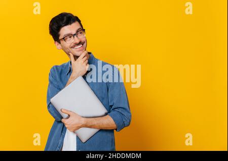 Positive smart guy in glasses and in a denim shirt, holding a laptop at hand, looks thoughtfully towards empty space aside touching his chin, smiling, isolated orange background,copy space Stock Photo