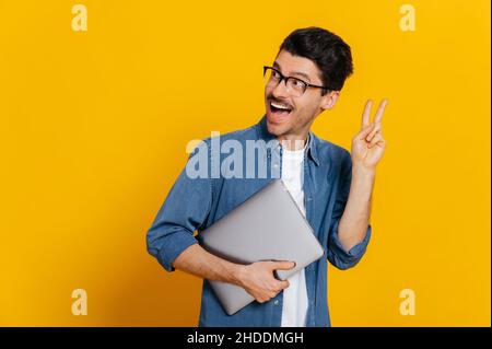 Friendly positive caucasian stylish handsome guy with glasses, freelancer or student, holds a laptop in hand, shows a peaceful gesture, stands on isolated orange background, looks back, smiling Stock Photo