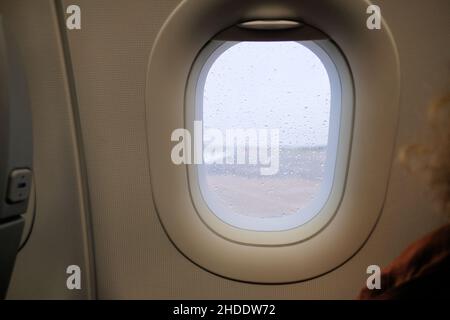 Airplane window during a storm with rain drops on window pane; selective focus on raindrops with blurry tarmac, wing, and gray exterior. Stock Photo