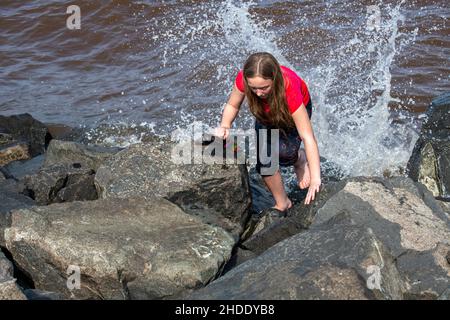 Duluth, Minnesota, Eleven-year-old girl climbing rocks on the shore of Lake Superior with huge wave crashing against the rocks. Stock Photo