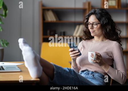 Young relaxed female employee enjoying break at work, using smartphone and drinking tea at workplace Stock Photo