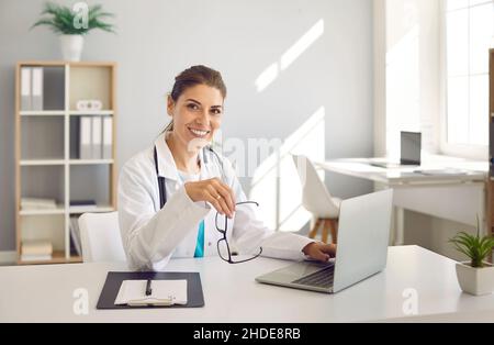 Happy female doctor sitting at working desk with laptop, looking at camera and smiling Stock Photo