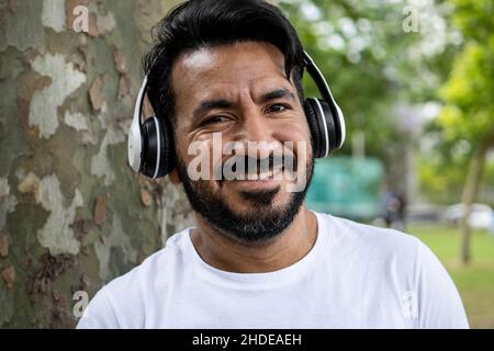 portrait Latin bearded man with headphones smiling at camera and listening to music in park Stock Photo