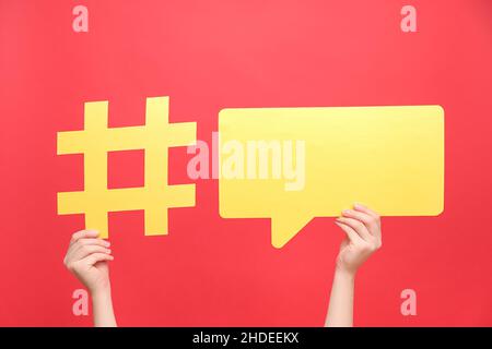 Female hands holding yellow paper hashtag symbol and speech bubble, hash sign of famous media content, isolated on red color background. Post viral we Stock Photo