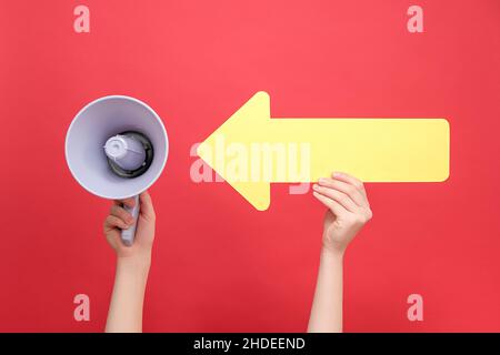 Close up of female hand holding megaphone pointing on big yellow arrow, isolated over red studio background wall with copy space for advertisement. Bu Stock Photo