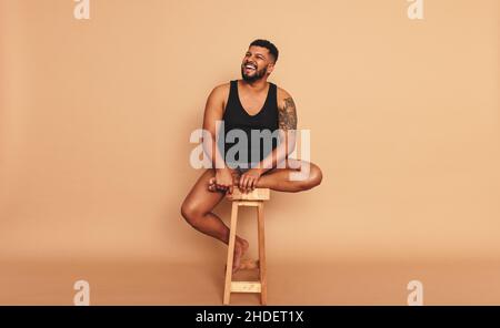 Tattooed man smiling cheerfully while sitting on a wooden chair in a studio. Body positive young man looking away while sitting alone. Self-confident Stock Photo