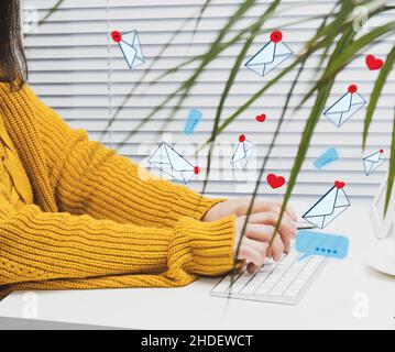 a woman sits at a white table and types on a wireless keyboard, letters and hearts are flying. Social media addiction, spam and online communication Stock Photo