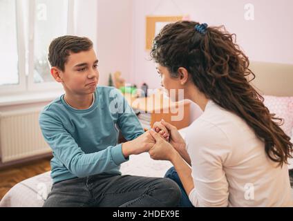 Mother holding kid for hands and having serious conversation but boy feels uncomfortable. Sitting on bed in sleeping room Stock Photo