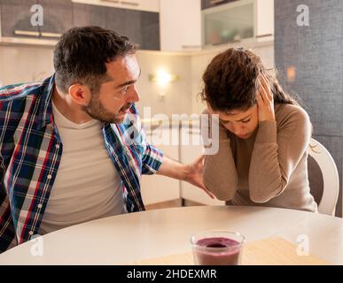 Husband and wife having argument at dining table at home. Man yelling with dominant posture and woman holding palms on ears and feeling frustrated Stock Photo