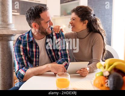 Happy couple sitting at dining table holding tablet and laughing Stock Photo