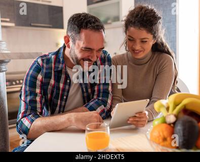 Happy couple sitting at dining table holding tablet and laughing Stock Photo