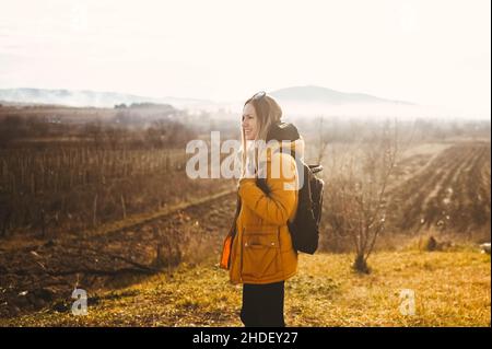 Young happy backpacker woman tourist traveling alone and posing at dawn with fog on background of beautiful nature with mountains in autumn. Blonde smiling lady in yellow down jacket Stock Photo