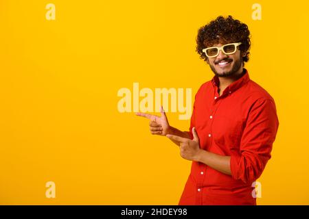 Portrait of pleasant looking bearded man wearing sunglasses pointing finger aside at copy space, showing area for advertisement. Indoor studio shot isolated on orange background Stock Photo