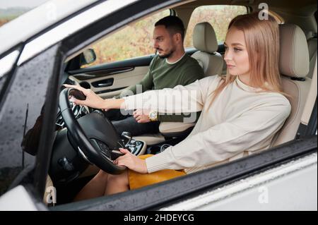 Side view of confident business blonde woman looking ahead, putting her hands on steering wheel of car. Successful lady driver and male passenger in front seat traveling in comfortable auto. Stock Photo