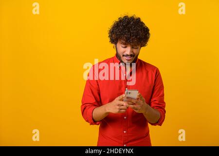 Waist up view of happy handsome blogger young adult man standing, using smartphone and texting message with smile. Indoor, isolated, studio shot, orange background, copy space Stock Photo