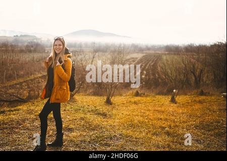 Young happy backpacker woman tourist traveling alone and posing at dawn with fog on background of beautiful nature with mountains in autumn. Blonde smiling lady in yellow down jacket Stock Photo