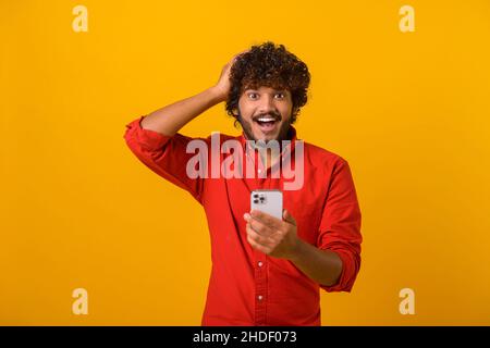 Curly brunette man with beard expressing amazement and holding head with his hands, while using cellphone, with open mouth and shocked big eyes. Indoor studio shot isolated on orange background Stock Photo