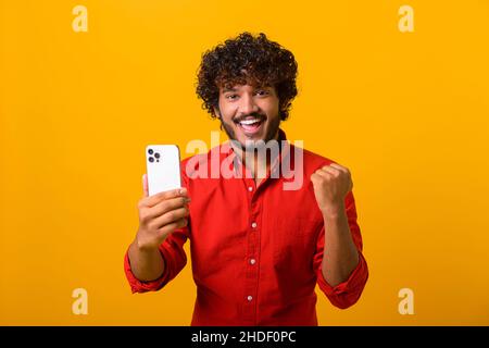 Happy satisfied man holding smartphone and smiling making yes gesture, celebrating online lottery or giveaway victory. Indoor studio shot isolated on orange background Stock Photo
