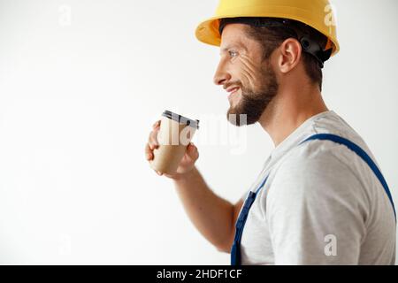Side view portrait of smiling handyman in uniform and hard hat drinking coffee during a break while standing indoors Stock Photo