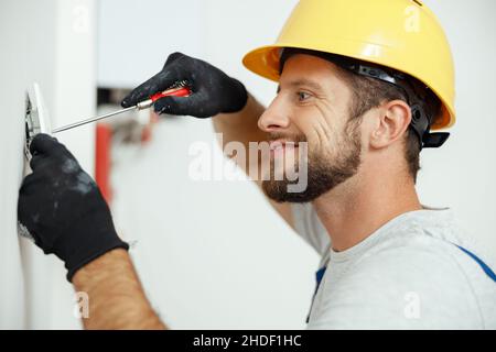 Smiling professional electrician in uniform replacing a socket in apartment after renovation work Stock Photo