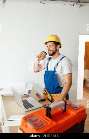 Relaxed handyman in uniform and hard hat drinking coffee during a break while doing renovation work indoors Stock Photo