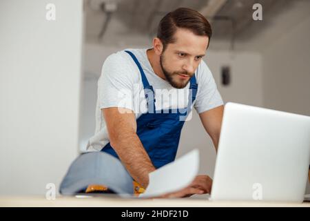 Focused professional handyman in workwear leaning on hands and looking at the screen of a laptop while standing indoors Stock Photo