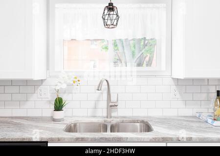 Kitchen sink detail shot with a marble countertop, white cabinets, stainless steel faucet and sink, and a light hanging in front of the window. Stock Photo