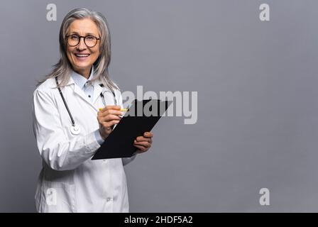 Senior doctor in white medical gown with stethoscope on shoulders taking notes standing with clipboard isolated on gray. Mature physician therapist writing down treatment plan, writes a prescription Stock Photo