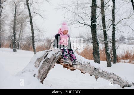 Distant portrait photo of female Russian kid sitting on tree log and smiling wearing pink winter clothes in forest. Astonishing background full of Stock Photo