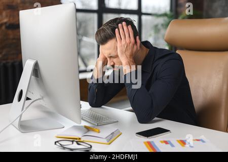 Tired caucasian guy sitting at the table at the office. Frustrated pensive man worried about work issues, covering head with hands in front of the computer Stock Photo