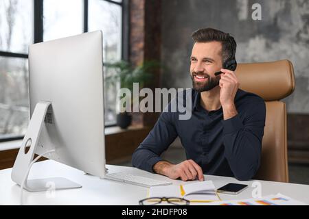 Office employees concept. Focused caucasian man wearing wireless headset is using laptop for online video conversation, talking with customers or coworkers online Stock Photo
