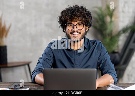 Portrait of smiling and friendly multi ethnic male freelancer in glasses using laptop and looks at the camera, indian latin curly guy in smart casual shirt typing, working remotely, develops software Stock Photo
