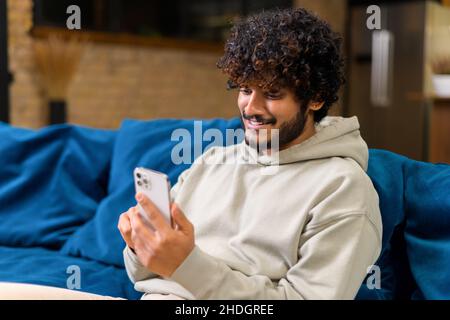 Smiling young curly Indian guy using smartphone sitting on the couch at home. Ethnic male in casual wear reading news online, messaging on the phone. Side view Stock Photo