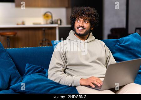 Cheerful Indian man wearing casual hoodie sitting on the sofa and using laptop at home. Inspired glad ethnic male entrepreneur typing on keyboard and looks aside with pleasure smile Stock Photo