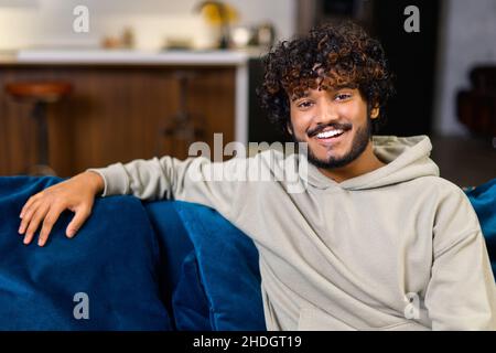 Portrait of carefree young Indian guy sitting on the sofa and looking at the camera, serene curly ethnic man in casual hoodie in relaxed atmosphere spending leisure at home Stock Photo